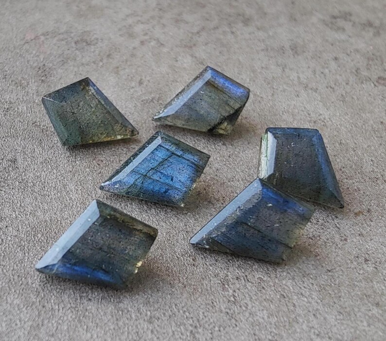 May include: Six trapezoid-shaped labradorite gemstones are displayed on a grey surface. The stones exhibit a mix of blue, green, and grey hues, with an iridescent shimmer. Each stone is faceted.