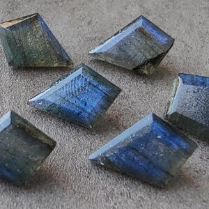 May include: Six faceted labradorite gemstones in shades of blue and grey. Each stone has a unique shape and cut, showcasing a play of colour. The gemstones are arranged on a neutral grey surface.