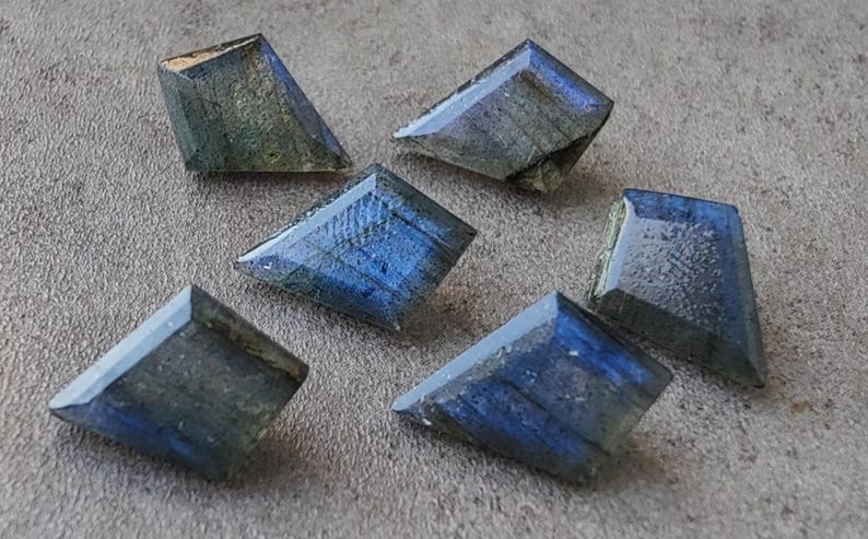 May include: Six faceted labradorite gemstones in shades of blue and grey. Each stone has a unique shape and cut, showcasing a play of colour. The gemstones are arranged on a neutral grey surface.