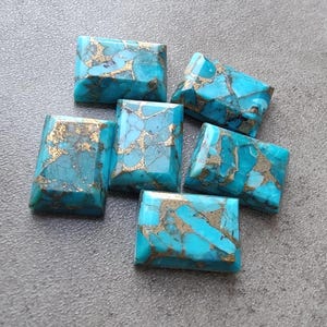 Könnte beinhalten: Sechs türkisfarbene Cabochons mit goldenen Flocken, in rechteckiger Form geschnitten. Die Steine sind in einer Gruppe auf einer grauen Oberfläche angeordnet.