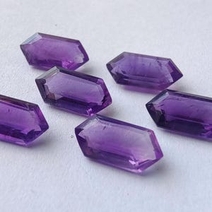 K&ouml;nnte beinhalten: Sechs facettierte Amethyst-Edelsteine ​​mit tiefvioletter Farbe sind auf einer wei&szlig;en Oberfl&auml;che angeordnet. Jeder Edelstein hat eine rechteckige Form mit abgewinkelten Enden, die die nat&uuml;rliche Farbe und Klarheit der Kristalle zeigen.