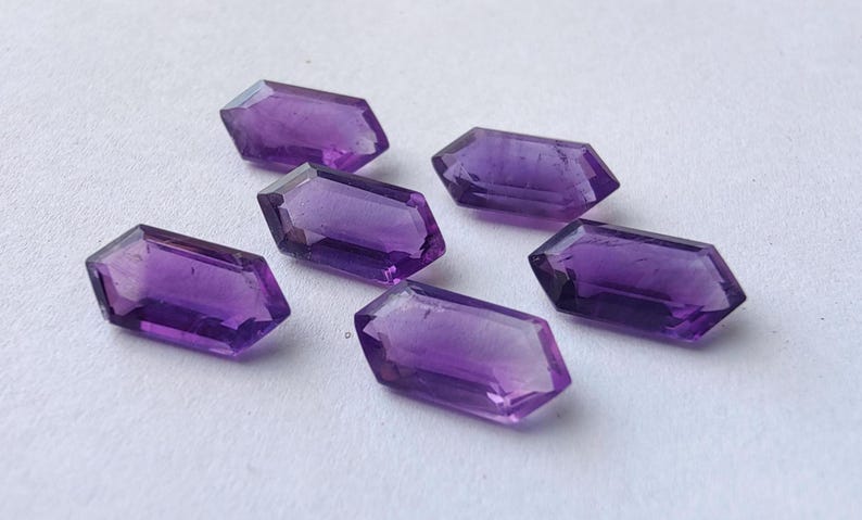 K&ouml;nnte beinhalten: Sechs facettierte Amethyst-Edelsteine ​​mit tiefvioletter Farbe sind auf einer wei&szlig;en Oberfl&auml;che angeordnet. Jeder Edelstein hat eine rechteckige Form mit abgewinkelten Enden, die die nat&uuml;rliche Farbe und Klarheit der Kristalle zeigen.