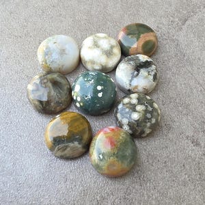May include: Nine polished gemstone cabochons in various colors and patterns, including white, green, brown, and red. The stones are round and have a smooth, glossy finish. The gemstones are arranged on a gray surface.