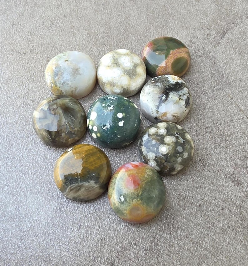 May include: Nine polished gemstone cabochons in various colors and patterns, including white, green, brown, and red. The stones are round and have a smooth, glossy finish. The gemstones are arranged on a gray surface.