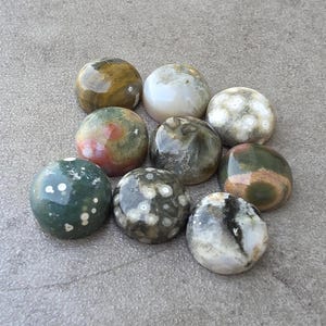 May include: A collection of nine polished, round gemstones in various colors, including green, brown, white, and red. The stones have unique patterns and textures, with some featuring speckled or marbled designs. The gemstones are arranged on a gray surface.
