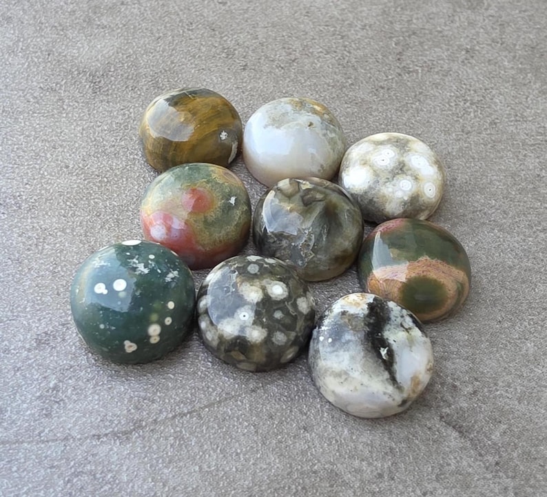 May include: A collection of nine polished, round gemstones in various colors, including green, brown, white, and red. The stones have unique patterns and textures, with some featuring speckled or marbled designs. The gemstones are arranged on a gray surface.