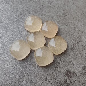 May include: Six translucent, heart-shaped gemstones with a pale yellow hue are arranged on a gray surface. The stones have a smooth, polished appearance, reflecting light. The gemstones are likely used for jewelry or crafts.