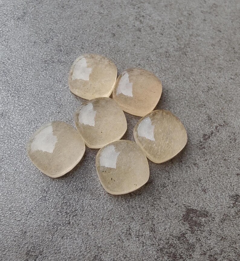 May include: Six translucent, heart-shaped gemstones with a pale yellow hue are arranged on a gray surface. The stones have a smooth, polished appearance, reflecting light. The gemstones are likely used for jewelry or crafts.