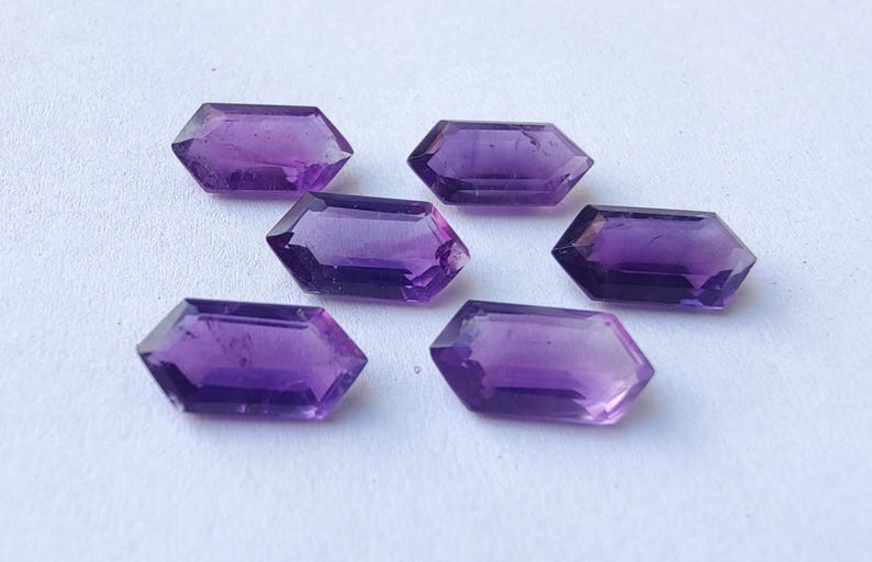 K&ouml;nnte beinhalten: Sechs facettierte, sechseckige Amethyst-Edelsteine. Die Edelsteine haben eine tiefviolette Farbe und sind auf einer wei&szlig;en Oberfl&auml;che angeordnet. Die Facetten fangen das Licht ein und erzeugen einen funkelnden Effekt. Geeignet f&uuml;r die Schmuckherstellung.