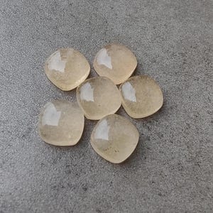 May include: Six translucent, square-cut gemstones with rounded corners are arranged on a gray surface. The stones have a pale yellow-brown hue and reflect light, creating highlights. The gemstones are likely for jewelry making or crafting.