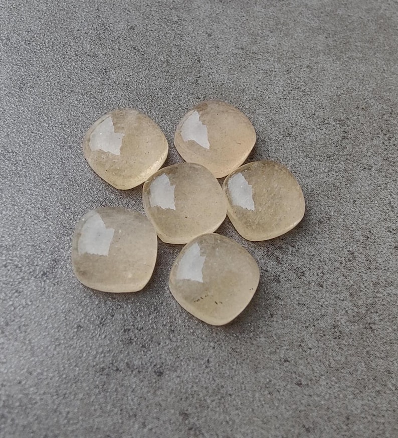 May include: Six translucent, square-cut gemstones with rounded corners are arranged on a gray surface. The stones have a pale yellow-brown hue and reflect light, creating highlights. The gemstones are likely for jewelry making or crafting.