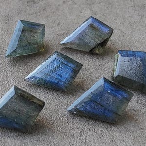 May include: Six trapezoid-shaped labradorite gemstones with a shimmering display of blue, grey, and green hues. The polished stones are displayed on a textured grey surface. Each stone has a faceted design.