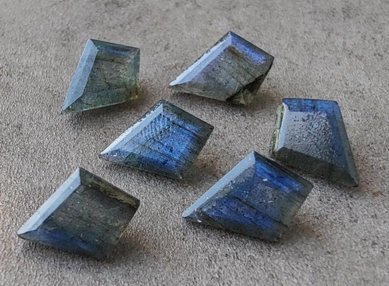 May include: Six trapezoid-shaped labradorite gemstones with a shimmering display of blue, grey, and green hues. The polished stones are displayed on a textured grey surface. Each stone has a faceted design.