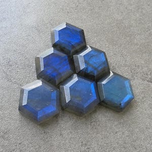 May include: Six hexagonal labradorite gemstones with a blue and gray color scheme. The gemstones are arranged in a stacked pattern, showcasing their polished facets and iridescent qualities. The stones are set against a gray background.