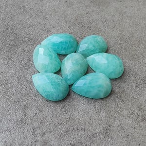 Op de afbeelding: Zeven gefacetteerde, druppelvormige amazoniet edelstenen in verschillende maten zijn gerangschikt op een grijze ondergrond. De edelstenen zijn licht turquoise van kleur met een subtiel, gevlekt patroon en een gepolijst oppervlak.