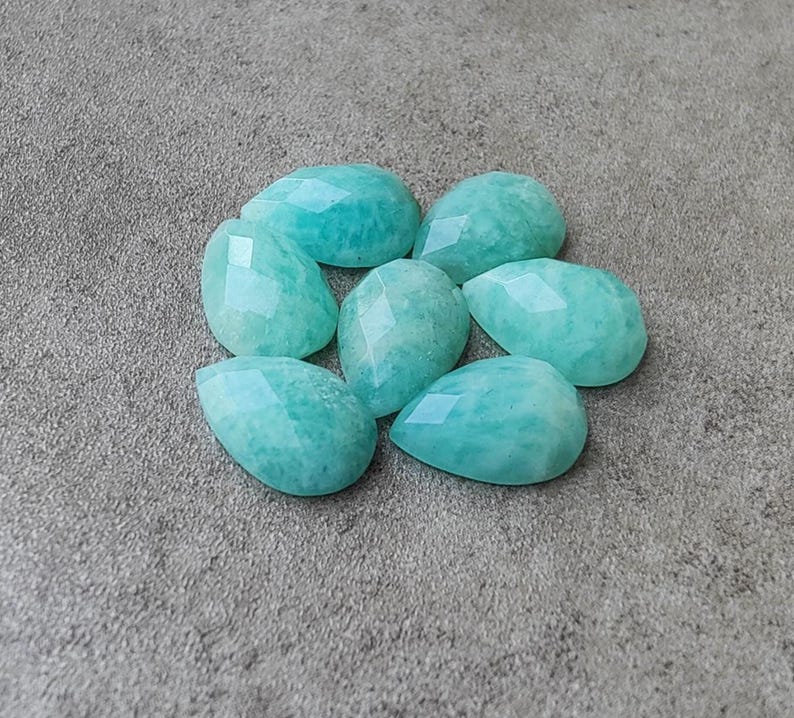 Op de afbeelding: Zeven gefacetteerde, druppelvormige amazoniet edelstenen in verschillende maten zijn gerangschikt op een grijze ondergrond. De edelstenen zijn licht turquoise van kleur met een subtiel, gevlekt patroon en een gepolijst oppervlak.