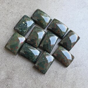 May include: A collection of rectangular gemstones with a polished, glossy finish. The stones are primarily dark green with red and brown inclusions. The arrangement of the stones is on a gray surface.