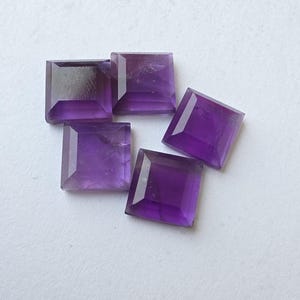 Peut inclure: Cinq pierres pr&eacute;cieuses d'am&eacute;thyste taill&eacute;es en carr&eacute;, de diff&eacute;rentes nuances de violet, sont dispos&eacute;es sur une surface blanche. Les pierres ont un design &agrave; facettes, captant la lumi&egrave;re et mettant en valeur leur couleur. Les pierres sont de taille et de forme similaires.