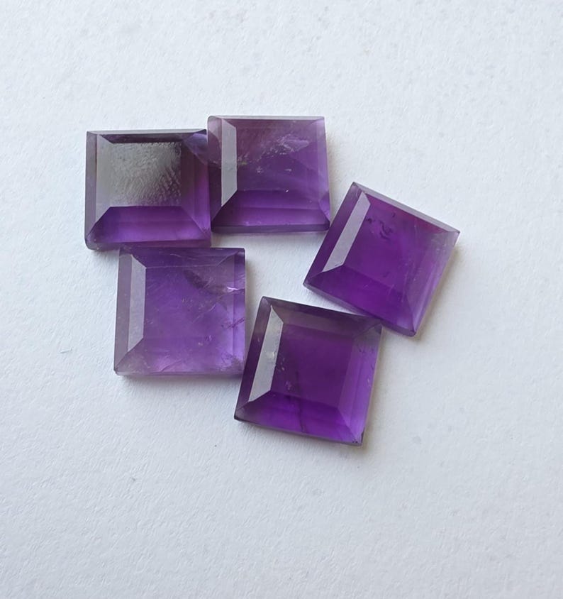 Peut inclure: Cinq pierres pr&eacute;cieuses d'am&eacute;thyste taill&eacute;es en carr&eacute;, de diff&eacute;rentes nuances de violet, sont dispos&eacute;es sur une surface blanche. Les pierres ont un design &agrave; facettes, captant la lumi&egrave;re et mettant en valeur leur couleur. Les pierres sont de taille et de forme similaires.