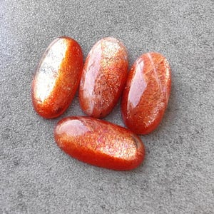 K&ouml;nnte beinhalten: Vier ovale Sonnenstein-Cabochons mit schimmerndem, irisierendem Finish. Die Steine sind eine Mischung aus Orange, Rot und Gold.