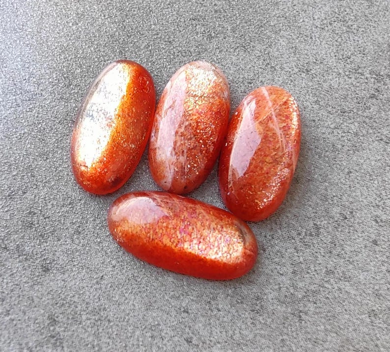 K&ouml;nnte beinhalten: Vier ovale Sonnenstein-Cabochons mit schimmerndem, irisierendem Finish. Die Steine sind eine Mischung aus Orange, Rot und Gold.