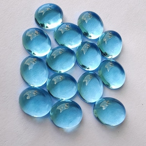 Puede incluir: Un grupo de 14 cabochones redondos de vidrio facetado de color azul claro. Los cabochones son lisos y brillantes, y están dispuestos en un grupo suelto.