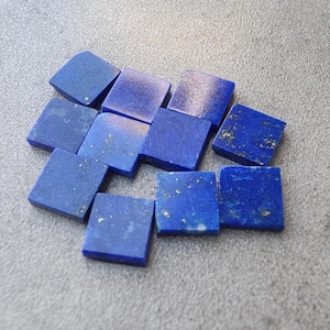 Könnte beinhalten: Eine Sammlung quadratischer Lapislazuli-Edelsteine. Die Steine sind tiefblau mit goldenen und weißen Flecken. Die Edelsteine sind auf einer grauen Oberfläche lose angeordnet.