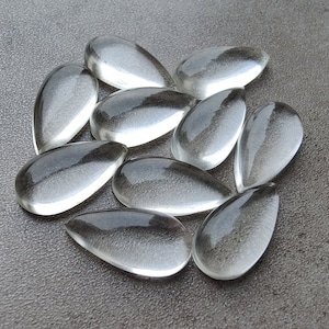 May include: A group of ten clear glass teardrop-shaped stones. The stones are arranged in a loose circle on a grey textured surface.