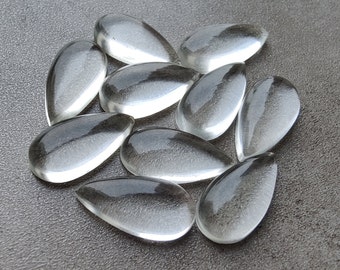 May include: A group of ten clear glass teardrop-shaped stones. The stones are arranged in a loose circle on a gray textured surface.