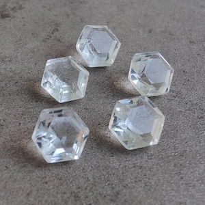 Puede incluir: Cinco piedras preciosas hexagonales transparentes y facetadas. Las piedras preciosas están dispuestas en semicírculo sobre una superficie gris.