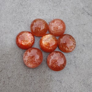 May include: Eight round, orange-red sunstone cabochons with a shimmery, glittery finish. The stones are arranged in a circular pattern on a gray background.