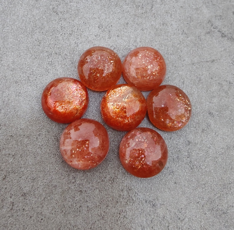 May include: Eight round, orange-red sunstone cabochons with a shimmery, glittery finish. The stones are arranged in a circular pattern on a gray background.