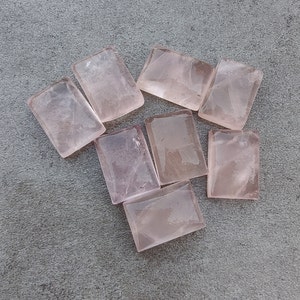 May include: Eight rectangular pink rose quartz stones, each with a different pattern of inclusions, arranged on a gray surface.