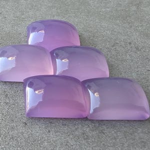 Pu&ograve; includere: Cinque pietre preziose lucide, color lavanda, con angoli arrotondati, sono disposte a forma di piramide. Le pietre hanno una superficie lucida e riflettente, e la luce cattura il loro aspetto liscio e traslucido. Lo sfondo &egrave; una superficie grigia strutturata.