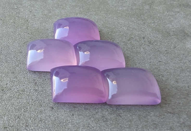 Pu&ograve; includere: Cinque pietre preziose lucide, color lavanda, con angoli arrotondati, sono disposte a forma di piramide. Le pietre hanno una superficie lucida e riflettente, e la luce cattura il loro aspetto liscio e traslucido. Lo sfondo &egrave; una superficie grigia strutturata.