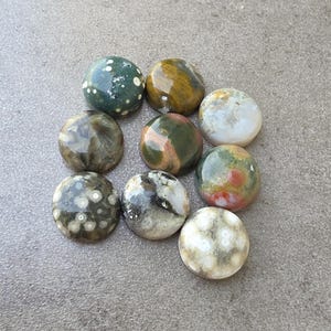 May include: Nine polished, round gemstones in various colors, including green, brown, white, and red. The stones have unique patterns and textures, with some featuring speckled or marbled designs. The gemstones are arranged on a gray surface.