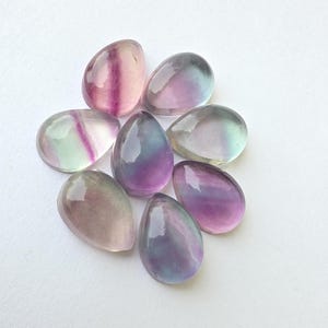 Può includere: Una collezione di otto pietre preziose di fluorite lucidata in varie tonalità di viola, verde e trasparente. Le pietre a forma di goccia sono disposte su una superficie bianca, mostrando la loro qualità traslucida e le variazioni di colore naturali.