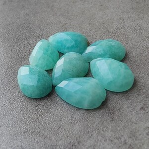 Op de afbeelding: Een verzameling van zeven gefacetteerde Amazoniet edelstenen in druppelvorm. De licht turquoise stenen zijn gepolijst en liggen op een grijze ondergrond.