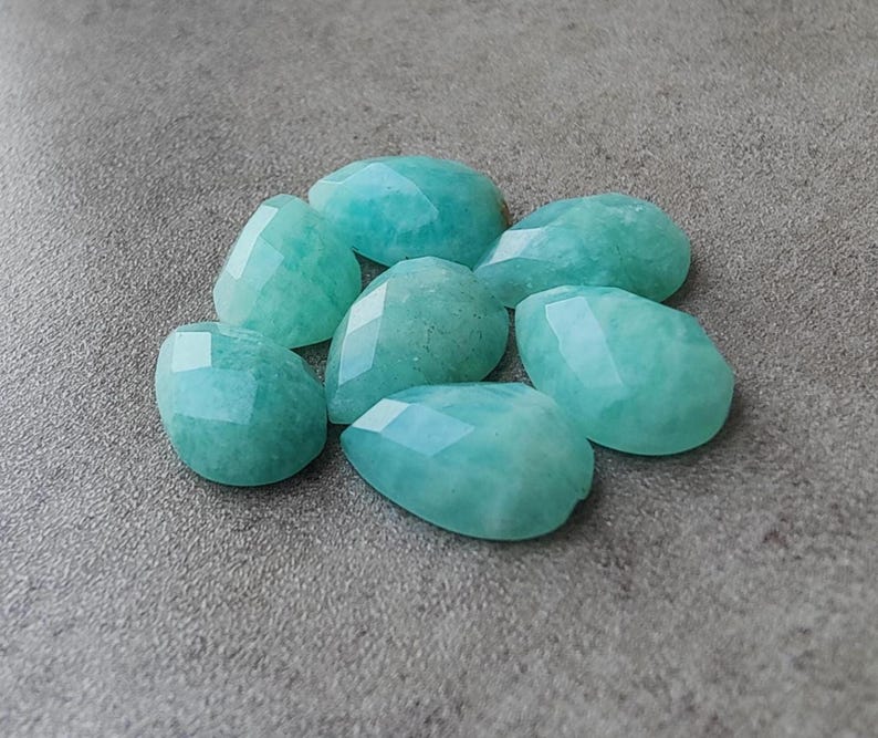 Op de afbeelding: Een verzameling van zeven gefacetteerde Amazoniet edelstenen in druppelvorm. De licht turquoise stenen zijn gepolijst en liggen op een grijze ondergrond.
