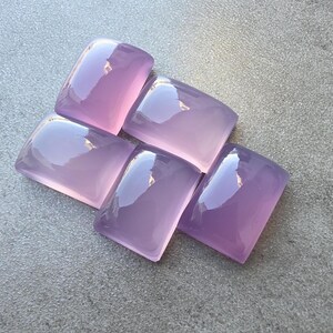 Pu&ograve; includere: Cinque pietre preziose lucide color lavanda con angoli arrotondati sono disposte su una superficie grigia. Le pietre hanno una superficie lucida e riflettente, che cattura la luce. Le gemme sono di un viola morbido e traslucido.