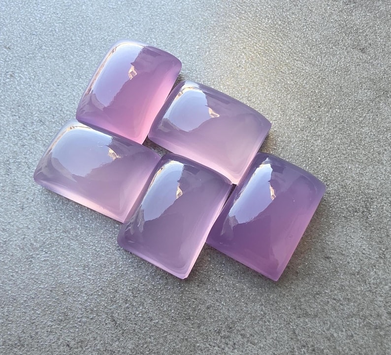 Pu&ograve; includere: Cinque pietre preziose lucide color lavanda con angoli arrotondati sono disposte su una superficie grigia. Le pietre hanno una superficie lucida e riflettente, che cattura la luce. Le gemme sono di un viola morbido e traslucido.