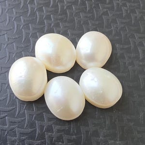 Op de afbeelding: Zes cr&egrave;mekleurige, ovale parels met een parelmoerglans liggen op een donkere, getextureerde ondergrond. De gladde, afgeronde vormen van de parels worden benadrukt. Parels zijn een klassiek element in sieraden.