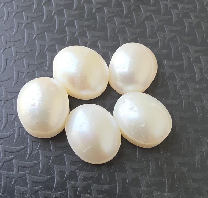 Op de afbeelding: Zes cr&egrave;mekleurige, ovale parels met een parelmoerglans liggen op een donkere, getextureerde ondergrond. De gladde, afgeronde vormen van de parels worden benadrukt. Parels zijn een klassiek element in sieraden.
