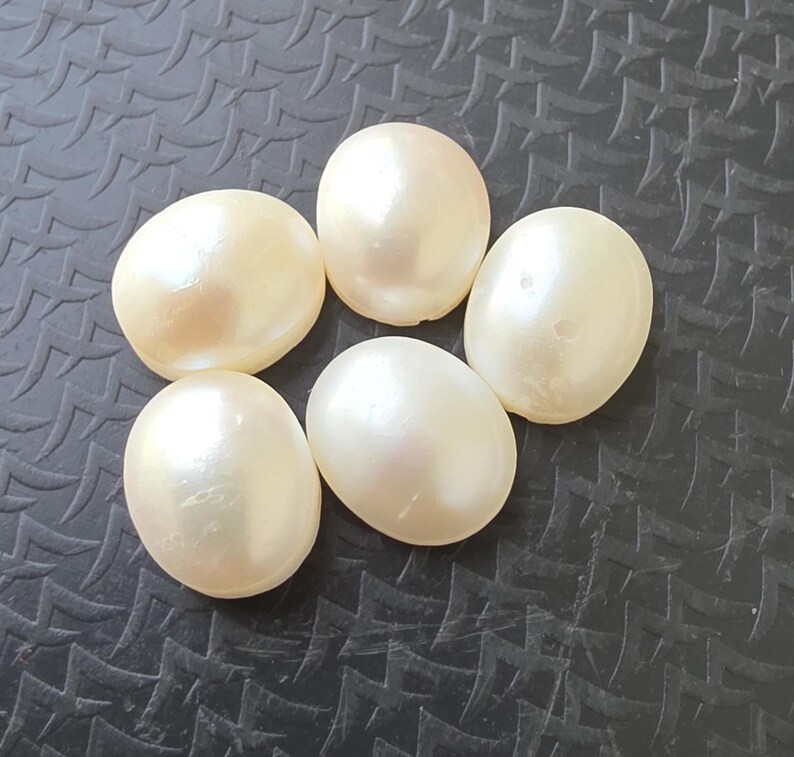 Op de afbeelding: Vijf glanzende, ovaalvormige parels in off-white op een donker, getextureerd oppervlak. De parels hebben een subtiele glans die het licht reflecteert. Geschikt voor het maken van sieraden. Elke parel is ongeveer 1,5 cm lang.