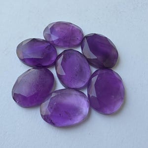 May include: Seven oval-shaped, faceted amethyst gemstones. The gemstones are a deep purple color and have a polished, reflective surface. The stones are arranged in a circular pattern against a white background.