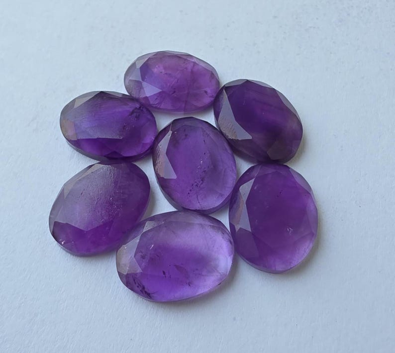 May include: Seven oval-shaped, faceted amethyst gemstones. The gemstones are a deep purple color and have a polished, reflective surface. The stones are arranged in a circular pattern against a white background.