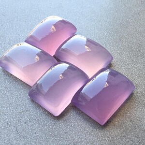 Pu&ograve; includere: Cinque cabochon di pietre preziose lucidate, color lavanda, disposti su una superficie grigia. Ogni cabochon ha una parte superiore arrotondata e una parte inferiore piatta, con una superficie liscia e riflettente. Le pietre sono di un viola morbido e traslucido.
