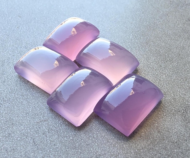 Pu&ograve; includere: Cinque cabochon di pietre preziose lucidate, color lavanda, disposti su una superficie grigia. Ogni cabochon ha una parte superiore arrotondata e una parte inferiore piatta, con una superficie liscia e riflettente. Le pietre sono di un viola morbido e traslucido.