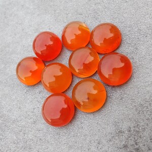May include: A group of eight orange carnelian cabochons, each with a smooth, polished surface. The stones are arranged in a circle on a gray background.