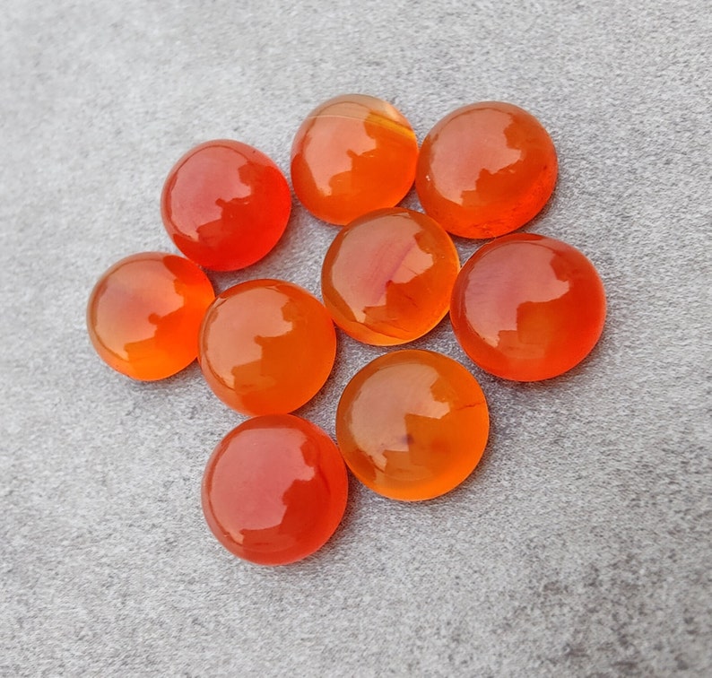 May include: A group of eight orange carnelian cabochons, each with a smooth, polished surface. The stones are arranged in a circle on a gray background.